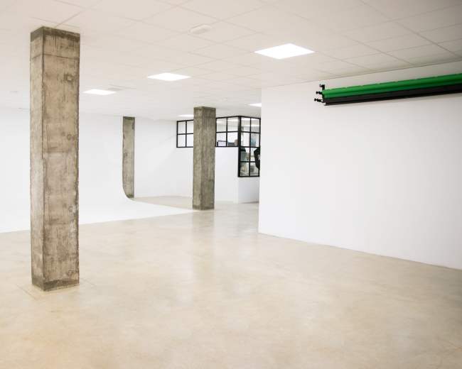 The image shows an empty studio space with concrete pillars, white walls, and a light-colored floor.