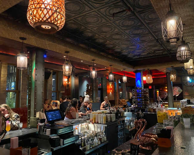 The image shows a lively bar interior with decorative pendant lighting, a busy counter with various drinks, and patrons engaged in conversation.