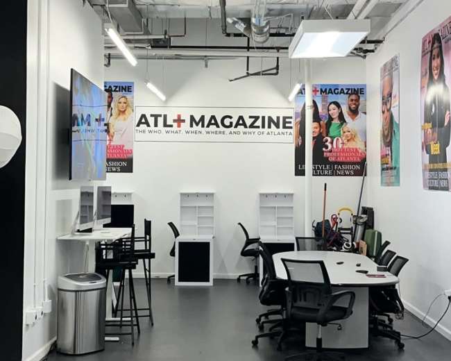 The image shows a modern office space with a conference table, black chairs, and promotional magazine covers displayed on the walls.