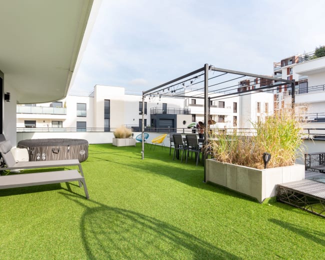 The image shows a rooftop terrace featuring artificial grass, lounge chairs, a hot tub, and a dining area with a pergola.