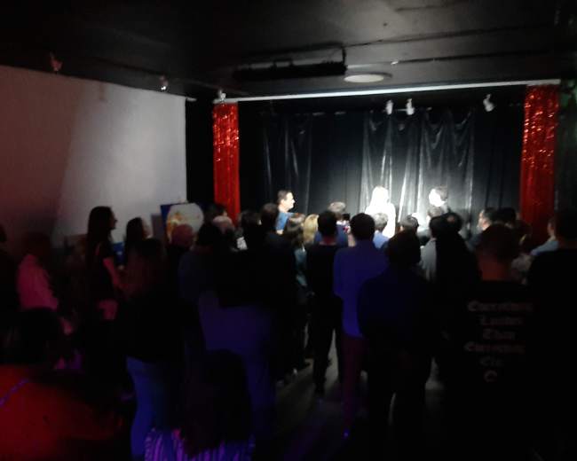 A crowd of people watches a performance on stage in a dimly lit venue adorned with red curtains.