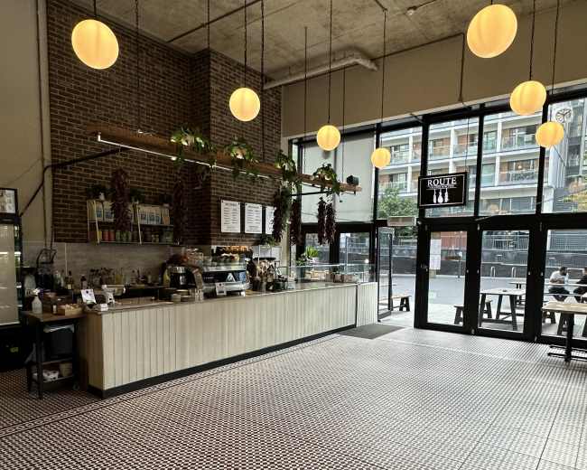The image shows a spacious cafe with a patterned tile floor, a long counter stocked with food and drinks, and large windows allowing natural light to enter.