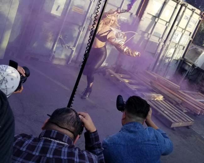 A model poses amidst purple smoke while photographers capture the scene in an outdoor setting with shipping containers in the background.