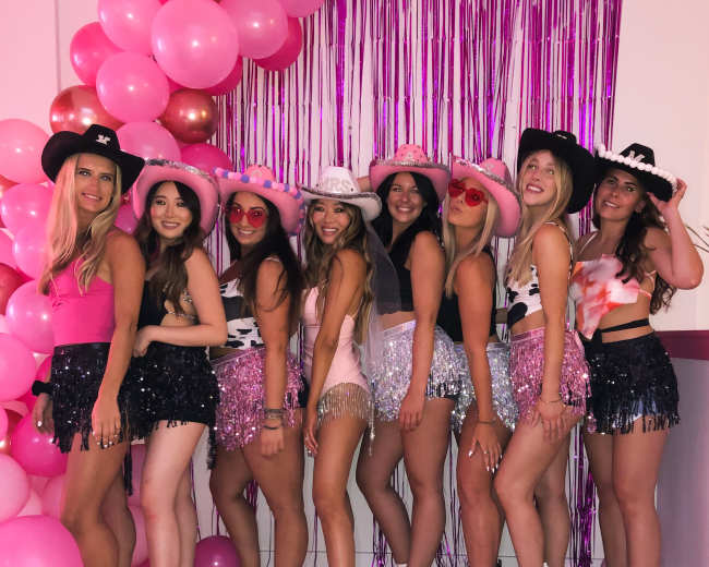 A group of eight women in glittery shorts and hats poses against a backdrop of pink balloons and shimmering curtains.