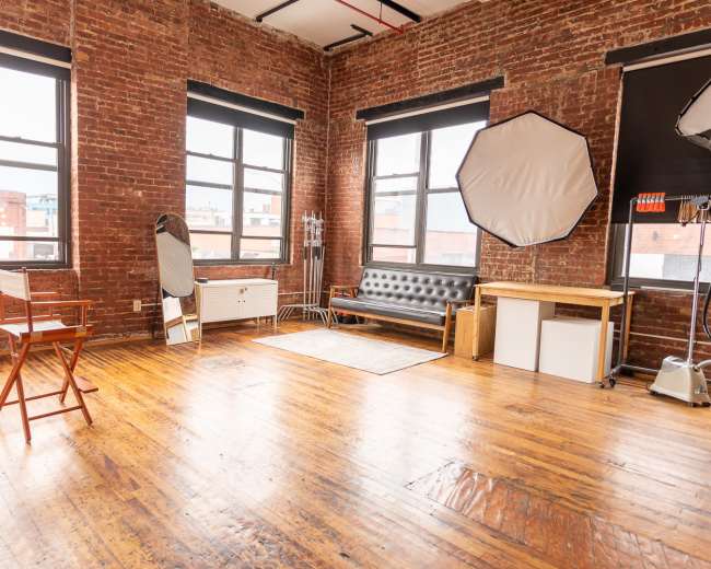 The image shows a spacious room with brick walls, large windows, wooden flooring, and minimal furniture including a chair, a couch, and a small table.