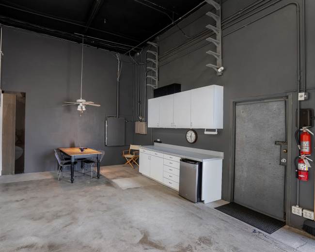 A spacious, minimally furnished room features a kitchenette with white cabinetry, a wooden table with chairs, and a gray wall backdrop.