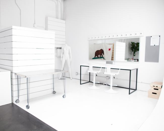 The image shows a modern, minimalist studio with white walls, a large mirror, a table on wheels, and a display area with slatted surfaces.