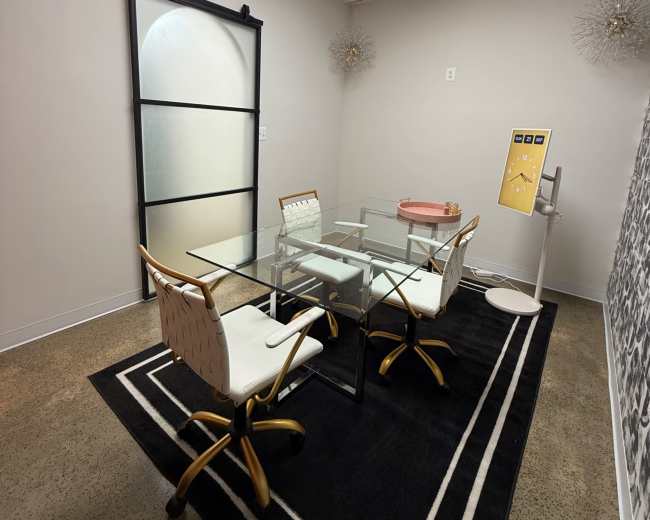 The image shows a small meeting room with a glass table, four chairs, a wall with decorative elements, and a sliding glass door.