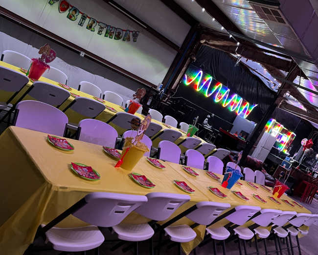 A decorated party space features a long table with yellow tablecloths, colorful plates, and a "Happy Birthday" banner.