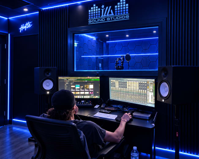 A person sits at a large workstation in a sound studio, focused on audio mixing software, surrounded by illuminated walls and sound equipment.