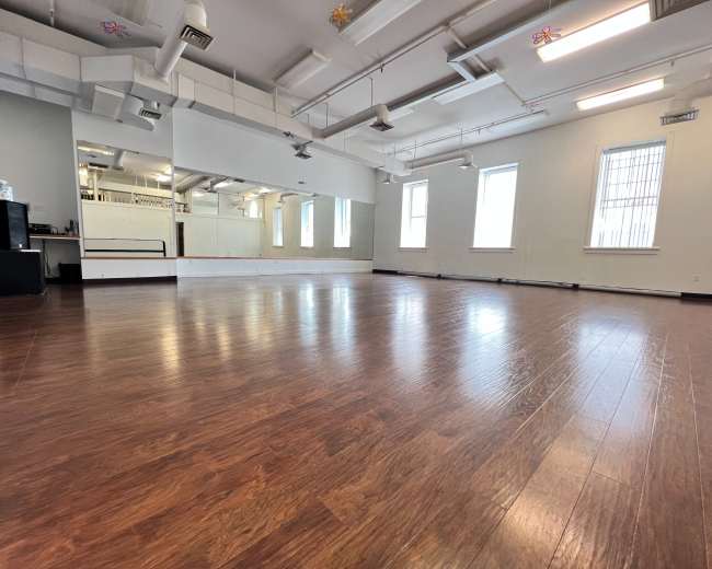 An empty dance studio with wooden flooring, large mirrors, and multiple windows allowing natural light to enter.