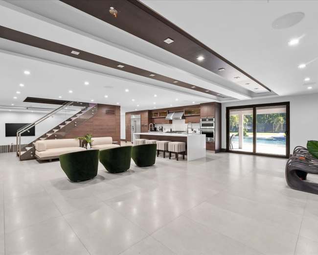 The image shows a modern, spacious living area with a kitchen, dining space, and large windows overlooking a pool.