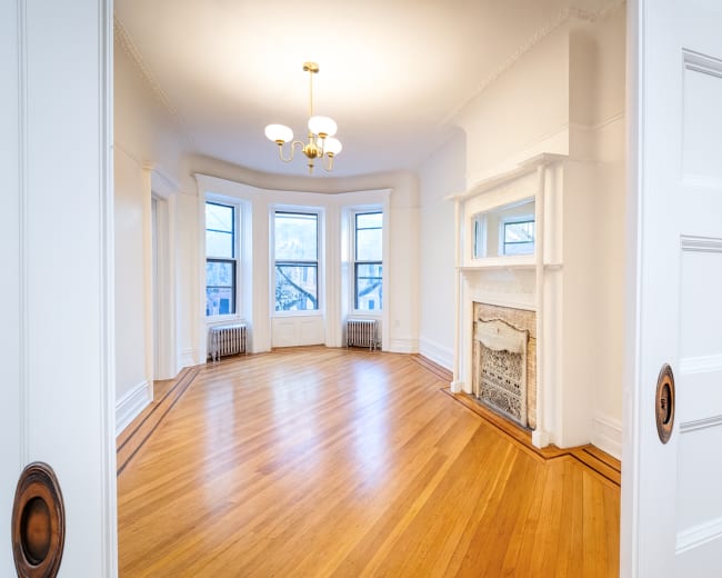 The image shows a bright, empty room with large windows, hardwood floors, and a decorative fireplace.
