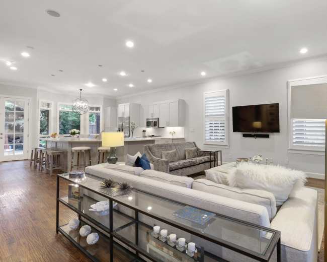 A modern living room with a light-colored sofa and a glass coffee table, adjacent to a kitchen area with white cabinetry and stainless steel appliances.