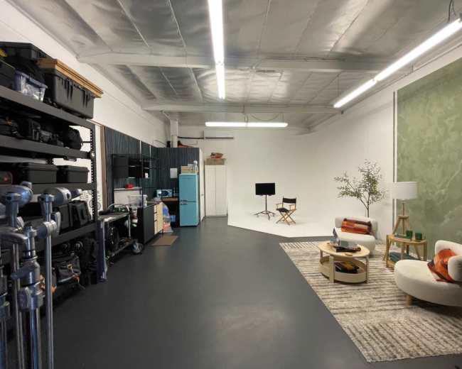 The image shows a spacious studio with shelves containing equipment on one side, a seating area with a lamp and coffee table, and a backdrop setup for video recording.