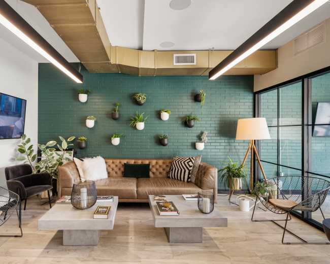 The image shows a modern lounge area with a brown leather sectional couch, concrete coffee tables, and a green wall adorned with potted plants.