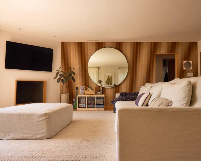 The image shows a modern living room featuring a large circular mirror, a sectional sofa, a coffee table, and a television mounted on the wall.