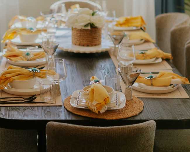 A neatly set dining table with decorative place settings, glasses, and a centerpiece of flowers is illuminated by natural light.