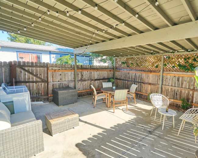 The image shows a covered outdoor patio area with several seating options, a small table, and a laundry area against a wooden fence.