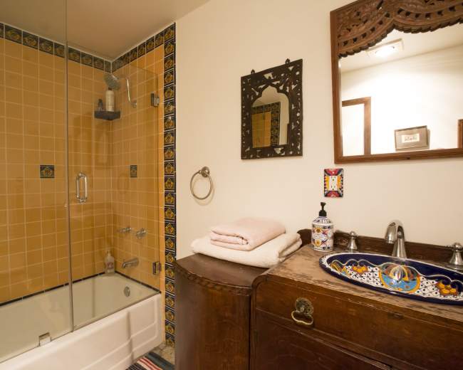 The image shows a bathroom featuring a glass-enclosed shower next to a bathtub, a wooden vanity with decorative tiles, and neatly stacked towels.