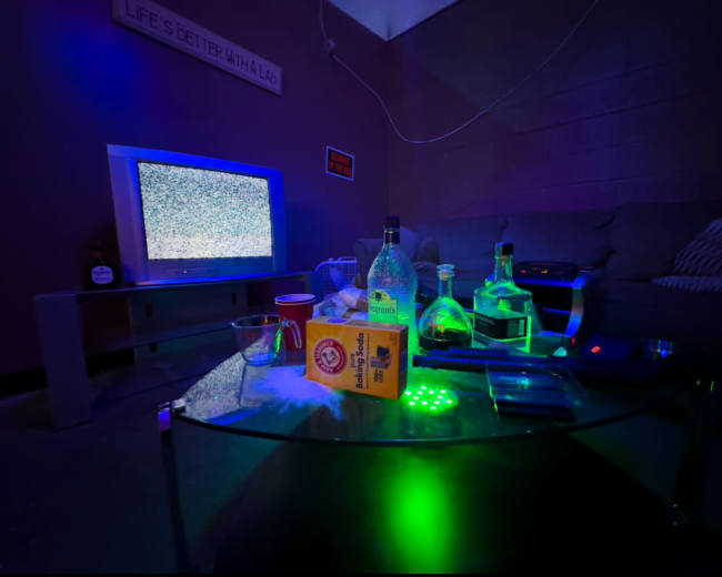 A living room with a vintage TV displaying static, a glass coffee table littered with bottles, a cup, and a box, and a couch in the background illuminated by green lighting.