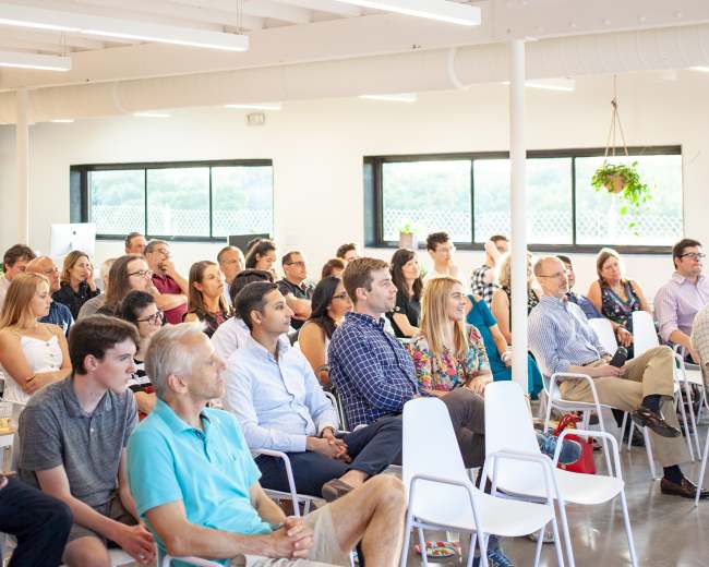 A diverse audience sits in a well-lit indoor space, attentively facing a speaker at the front.