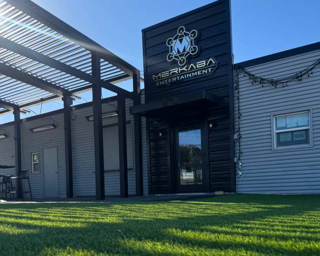 The image shows a modern building with a sign reading "Merkaba Entertainment," surrounded by a green lawn and shadowed by a pergola.