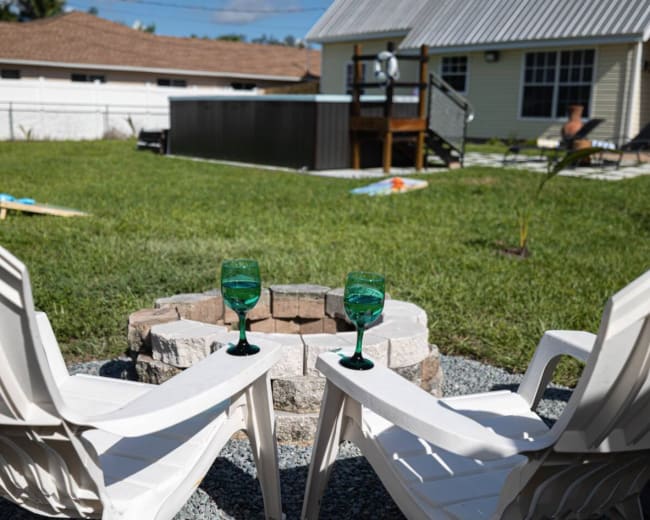 Two white plastic chairs are positioned around a circular stone fire pit, each holding a green goblet, with a pool and house visible in the background.