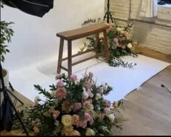 The scene features a wooden bench surrounded by large floral arrangements on a white backdrop in a studio setting.