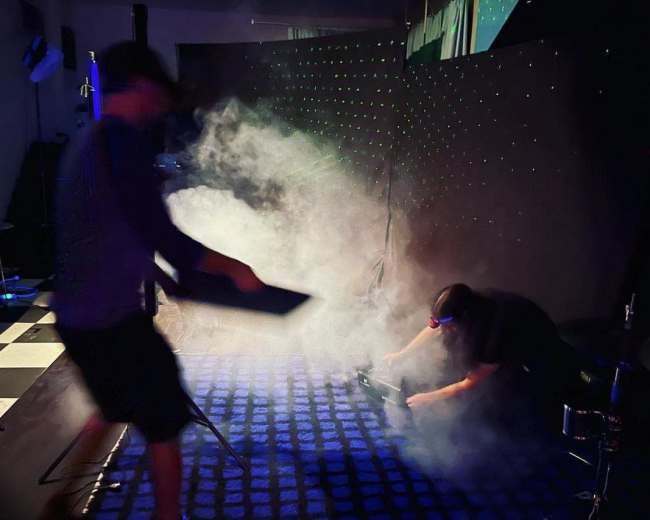 Two individuals are working in a smoky studio with a checkered floor, one adjusting equipment while the other is positioned on the ground.
