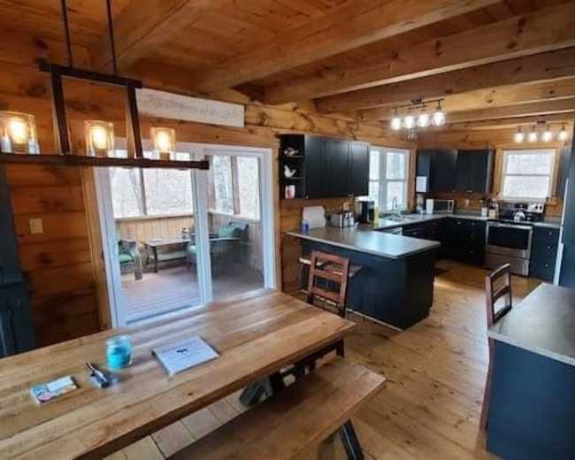 A rustic kitchen with wooden beams, dark cabinets, a central island, and a dining table, featuring large windows that overlook a deck.
