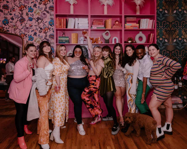 A group of women pose together in a colorful room adorned with floral wallpaper and a vibrant pink bookshelf.