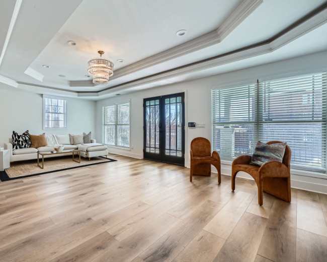 The image shows a spacious, well-lit living room featuring a large sofa, two chairs, a decorative area rug, and large windows with blinds.