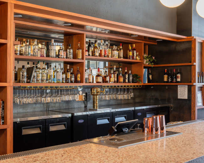 A bar with a wooden shelf filled with various bottles of liquor, glassware hanging beneath, and a polished countertop in front.