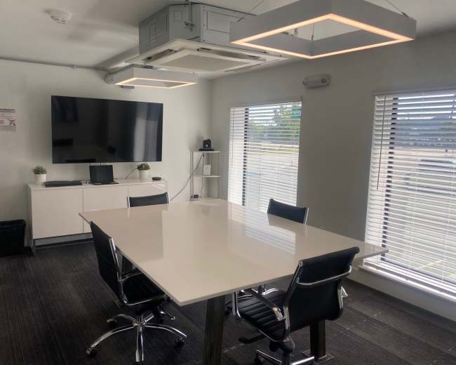 A modern conference room features a large white table surrounded by black chairs, a wall-mounted television, and window blinds.