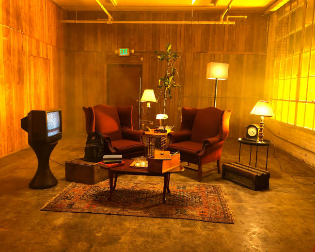 The scene features two red armchairs facing each other in a room with wooden walls, illuminated by warm lighting, accompanied by a vintage television, a side table with a lamp, a clock, and a potted plant.