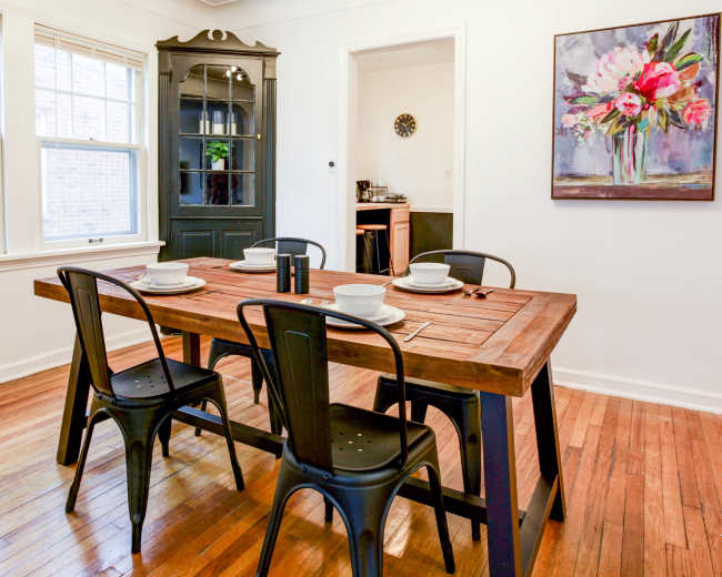 A wooden dining table with black chairs is set for four in a well-lit dining area featuring a black cabinet and a floral painting on the wall.