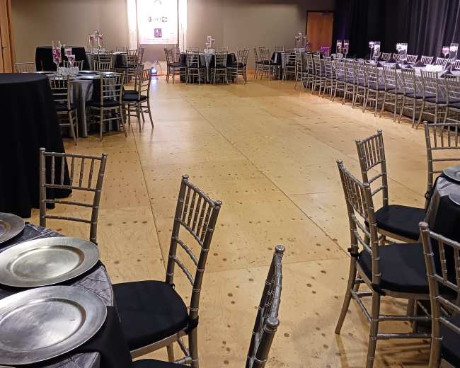 The image features a spacious event hall arranged with tables and chairs, illuminated by overhead lights and a backdrop for a formal gathering.