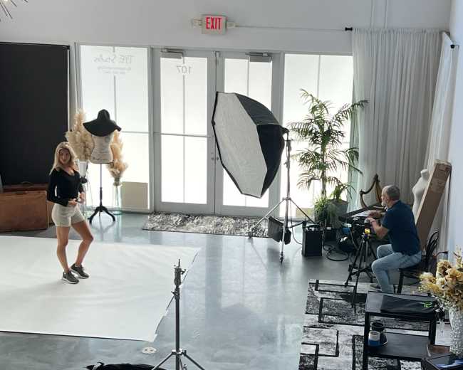 A model poses on a white backdrop while a photographer captures images from behind a camera in a studio setting.
