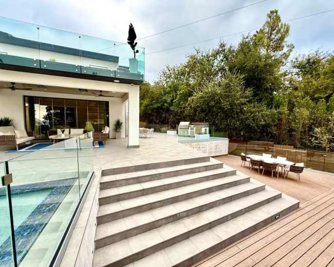 The image shows a modern outdoor space featuring a swimming pool, steps leading to a patio area, and a glass-enclosed building adjacent to lush greenery.