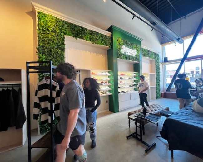 The image shows a clothing store interior with greenery on the walls, displaying various apparel items on shelves, and several customers browsing.