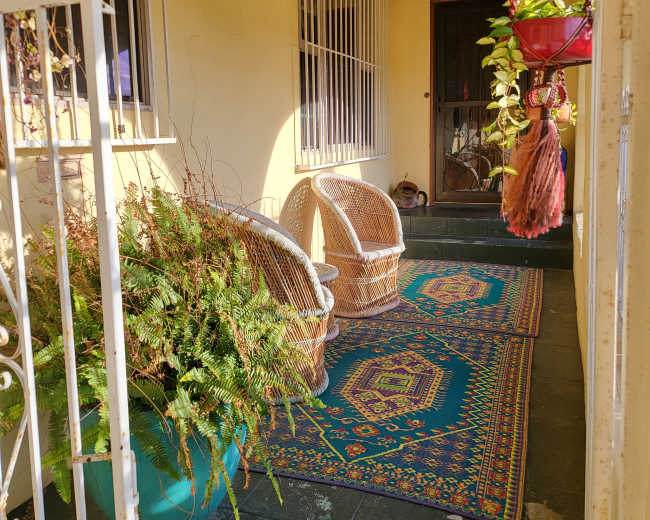 The image shows a porch area with two wicker chairs, a colorful area rug, hanging plants, and a decorative entrance door.