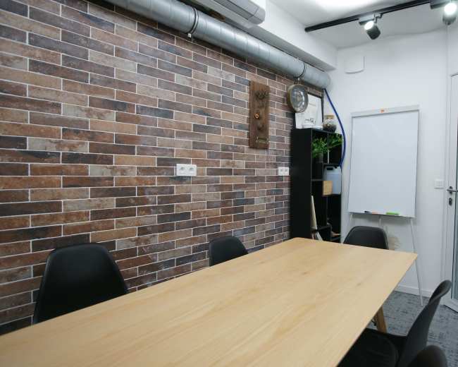 The image shows a small meeting room with a wooden table, black chairs, a brick-patterned wall, and a whiteboard on the side.