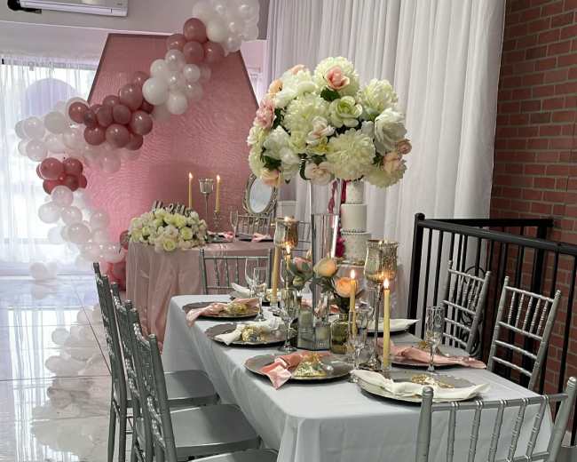 A decorated dining area features a long white table with elegant chairs, floral centerpieces, and candles, set against a backdrop of pink and white balloons.