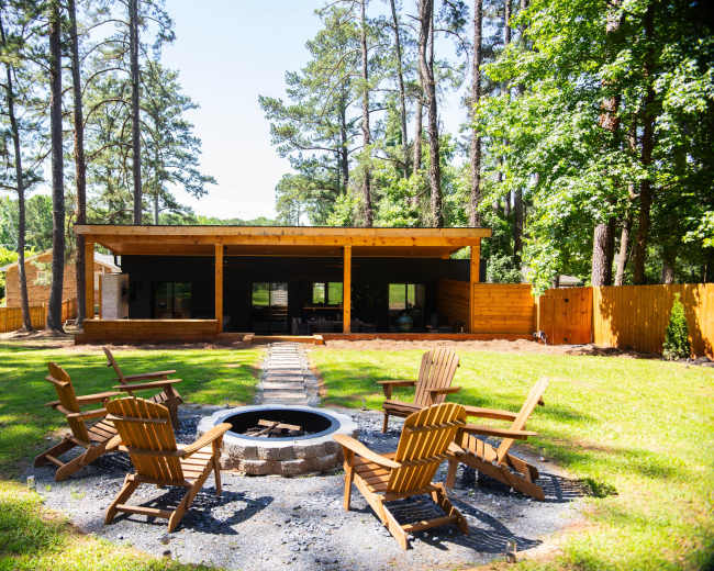 A circular arrangement of wooden chairs surrounds a stone fire pit in a green yard adjacent to a modern wooden structure and tall trees.