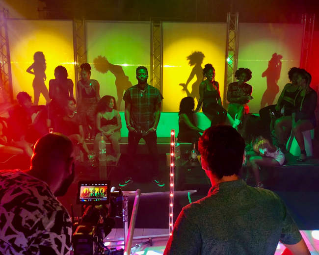 A group of people is gathered in a vibrant, illuminated setting, with some sitting on couches and others standing against colorful backdrops.