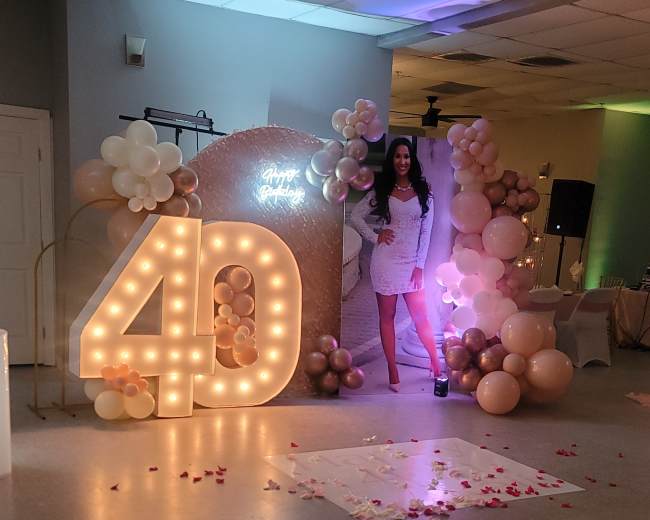 A woman stands next to a large illuminated "40" display, surrounded by decorative balloons and rose petals scattered on the floor.
