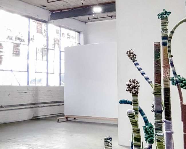 The image features an art gallery with an empty white wall and large windows allowing natural light, complemented by textile sculptures resembling plants in the foreground.