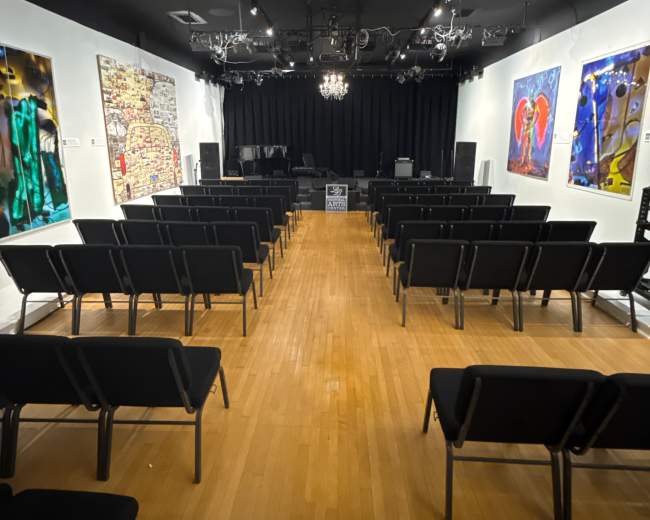 The image shows a performance space with rows of black chairs facing a stage, adorned with colorful artworks on the walls.