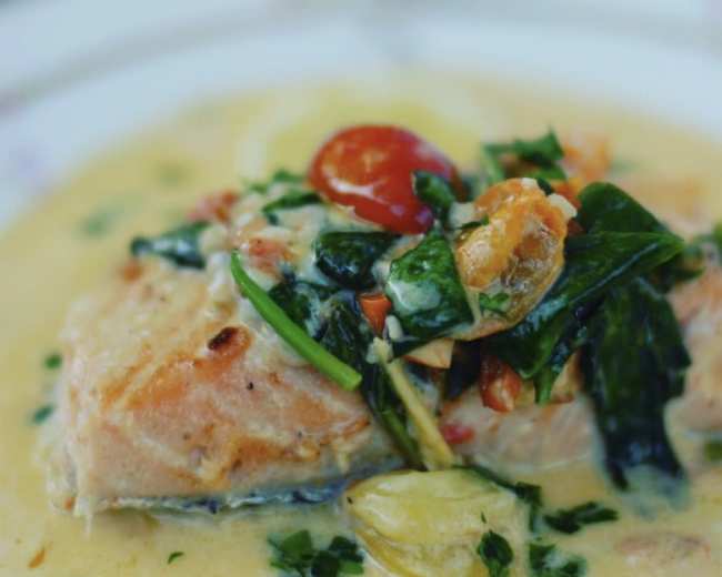 A plate of salmon topped with spinach, cherry tomatoes, and a creamy sauce is being held by a person.